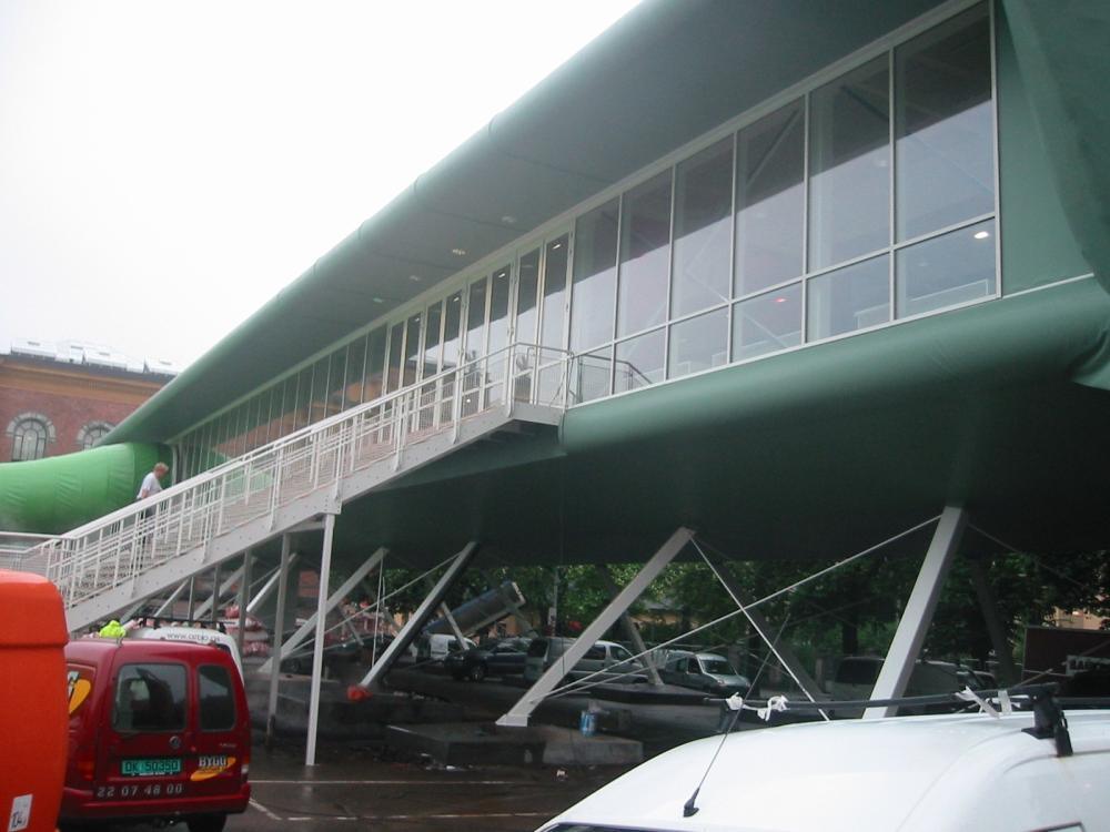 A building clad in green with a glazed walkway on steel legs